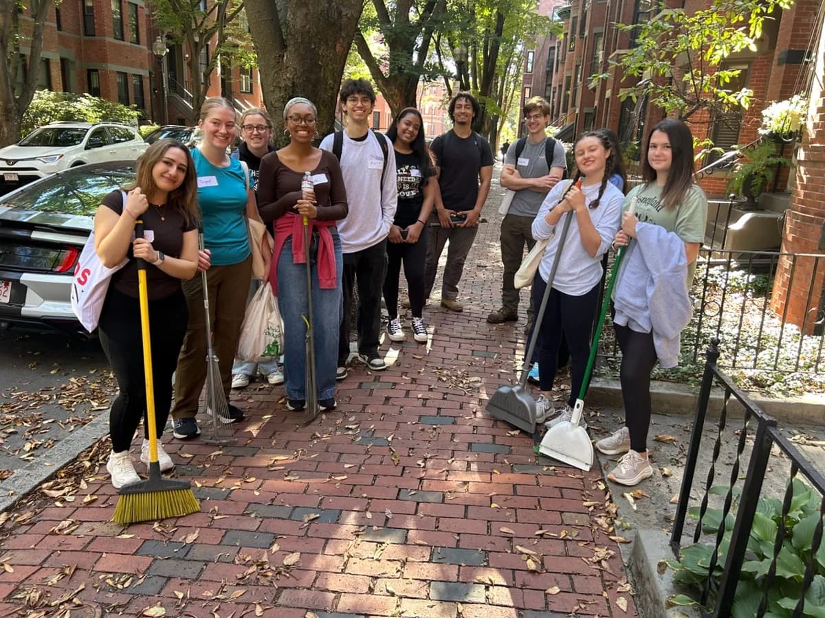 A group stands on a sidewalk holding brooms, a rake, and dustpans