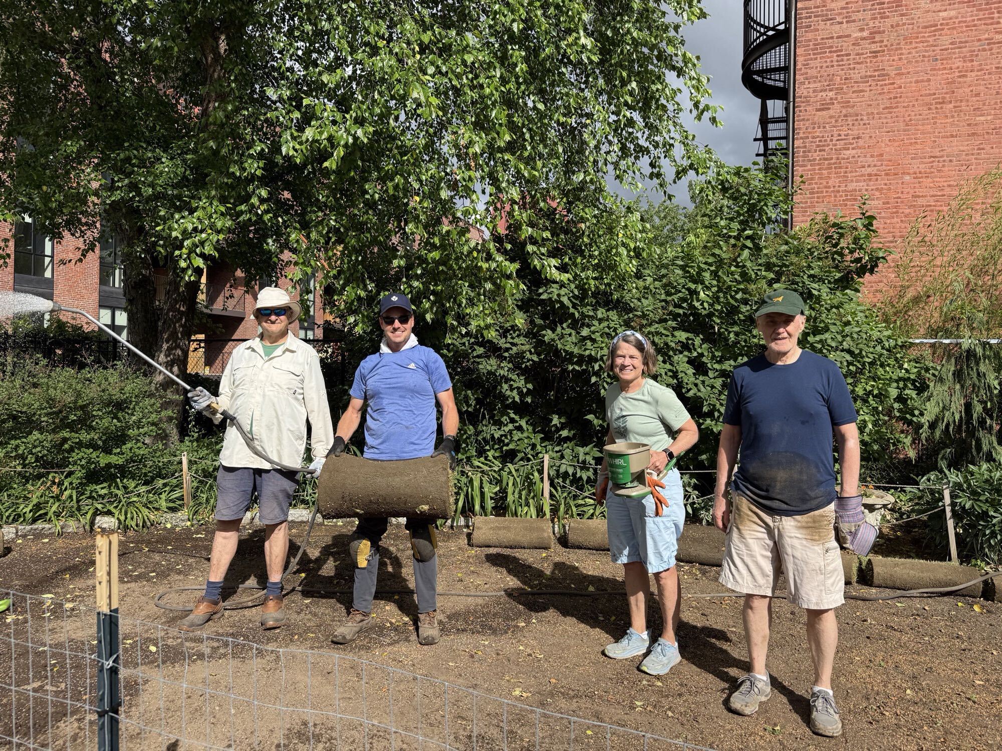 Four volunteers taking a break during sod installation, one holding a roll of sod