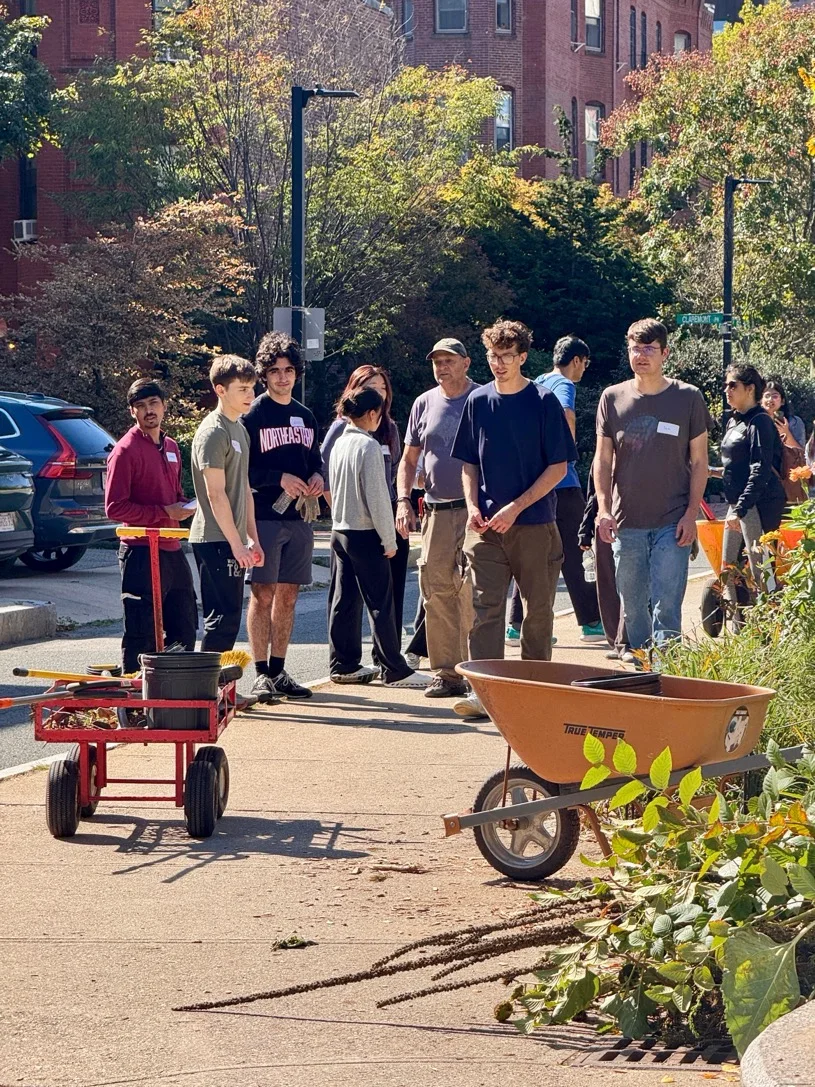 A group stands beside a wheelbarrow, a tool cart, and cut vegetation